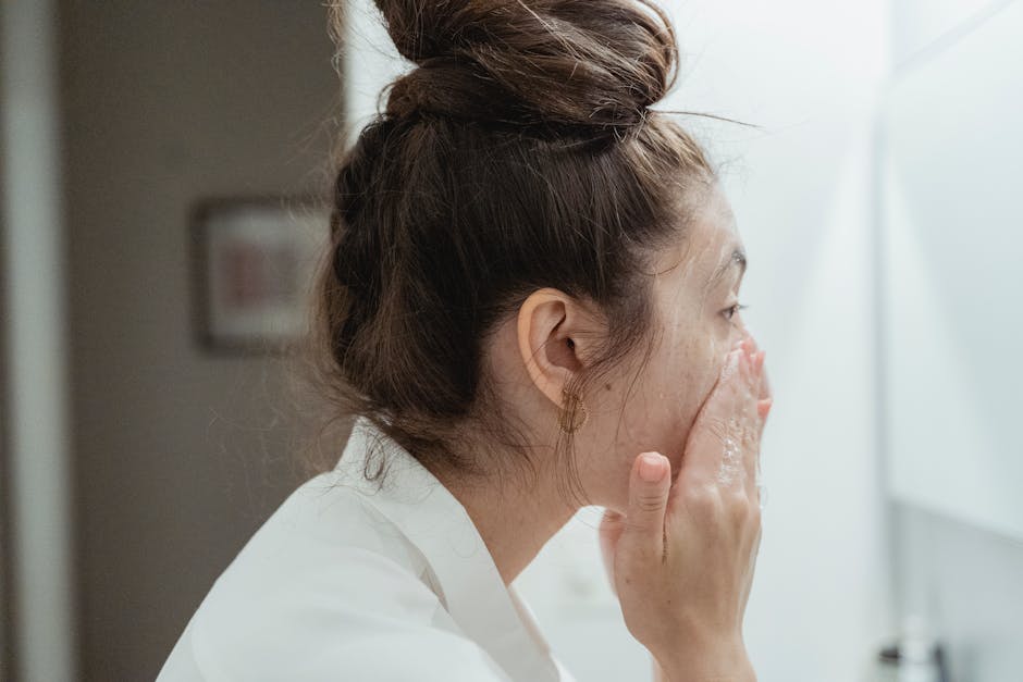 A young woman in a bathrobe follows her skincare routine, washing her face in a bathroom